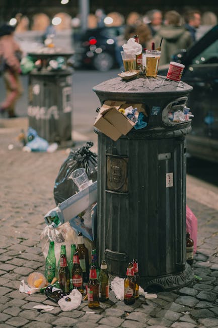 A black, cylindrical public trash bin with a textured surface located on a cobblestone pavement in an urban setting. The top of the bin is filled with various waste items, including crumpled paper cups, plastic cups with remnants of beverages, and discarded food wrappers. The side of the bin has visible slots for waste disposal, and around its base, there are multiple empty glass bottles, crumpled paper, and plastic waste scattered on the ground, some leaning against the bin and others lying on the pavement. Behind the bin, a grey metal trash receptacle with a partially open lid is visible, along with blurred figures of pedestrians and parked vehicles, suggesting a busy street scene. The lighting indicates an overcast day or late afternoon, with diffuse natural light illuminating the area. This scene illustrates issues related to improper rubbish disposal and highlights the importance of suitable waste management services such as those provided by Rubbish Removal Kensington, especially for areas experiencing high foot traffic and continuous waste generation. The cluttered environment emphasizes the need for effective rubbish collection and independent waste handling options within urban communities.