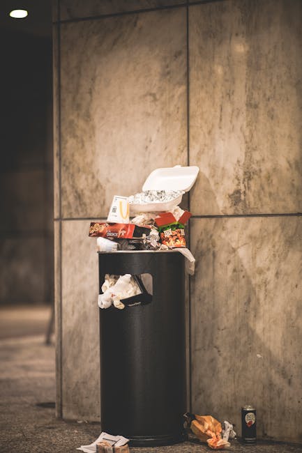A black cylindrical trash bin with its lid open, situated against a large, beige tiled wall in an indoor or underground public space. The bin is overflowing with various waste items, including food packaging, a paper container, and other miscellaneous rubbish. Some trash, such as a crumpled paper bag and an empty drink can, is scattered on the floor around the base of the bin, indicating it is overfilled. The environment appears clean but cluttered due to the excess waste. The lighting is neutral and even, highlighting the textures and colors of the waste and the smooth surface of the wall behind. This scene illustrates improper waste disposal, emphasizing the importance of responsible rubbish management and the potential need for additional waste collection services, such as those offered by Rubbish Removal Kensington for alternative waste handling and on-site clearance solutions in urban areas.