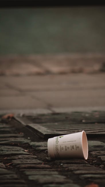 A black, cylindrical public trash bin with a textured surface located on a cobblestone pavement in an urban setting. The top of the bin is filled with various waste items, including crumpled paper cups, plastic cups with remnants of beverages, and discarded food wrappers. The side of the bin has visible slots for waste disposal, and around its base, there are multiple empty glass bottles, crumpled paper, and plastic waste scattered on the ground, some leaning against the bin and others lying on the pavement. Behind the bin, a grey metal trash receptacle with a partially open lid is visible, along with blurred figures of pedestrians and parked vehicles, suggesting a busy street scene. The lighting indicates an overcast day or late afternoon, with diffuse natural light illuminating the area. This scene illustrates issues related to improper rubbish disposal and highlights the importance of suitable waste management services such as those provided by Rubbish Removal Kensington, especially for areas experiencing high foot traffic and continuous waste generation. The cluttered environment emphasizes the need for effective rubbish collection and independent waste handling options within urban communities.