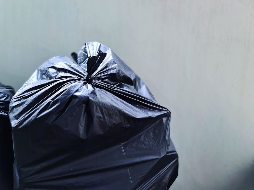 Close-up view of three large black plastic rubbish bags, tightly knotted at the top, sitting on a flat surface. The bags are made of shiny, durable plastic material with visible creases and folds. They appear to be filled with waste, causing the plastic to bulge and stretch. The background is plain and neutral, with a muted beige or light grey wall that provides minimal distraction. The lighting is soft and even, highlighting the glossy texture of the plastic bags. The scene suggests an indoor or outdoor location suitable for private waste collection, and the image aligns with the services of Rubbish Removal Kensington, focusing on the collection and disposal of waste materials outside of local authority rubbish collection. The composition emphasizes the practicality of managing rubbish through independent rubbish removal services, such as those offered by the company.
