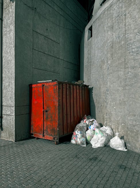 A large pile of black plastic rubbish bags, some of which are torn open revealing plastic bottles and paper waste, is stacked against a tiled wall in an outdoor urban area. The bags vary in size and appear to be filled with household waste. Behind the pile, the wall has graffiti with the word 'XEND' spray-painted in red. In the background, part of a modern building with reflective surfaces and a cloudy sky are visible, indicating an outdoor waste collection site or an area awaiting private rubbish removal. The scene illustrates unmanaged waste accumulation, highlighting the need for professional rubbish removal services like those offered by Rubbish Removal Kensington, which specialises in alternative waste handling to prevent such unsightly scenes in London neighborhoods.
