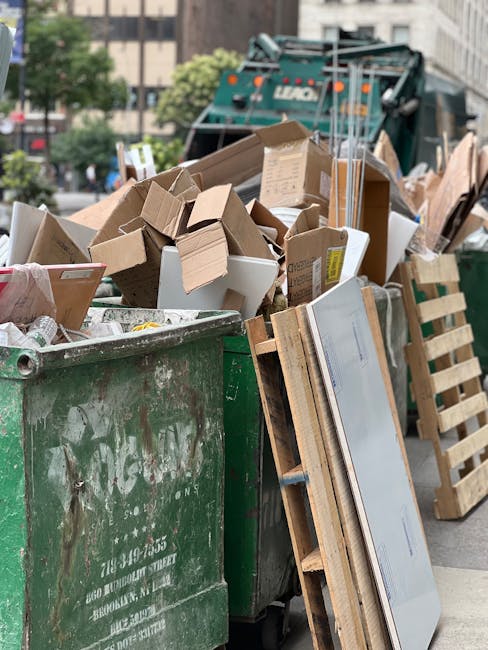 A large green waste bin filled with various types of discarded packaging, including cardboard boxes, some flattened and others partially intact, along with white polystyrene containers and packaging materials. The surface of the bin is weathered with visible dirt and scratches. Leaning against the bin are wooden pallets, some upright and others resting on the ground, which appear used and slightly worn. In the background, a green waste collection truck with the logo partially visible is parked on an urban street, surrounded by trees, residential buildings, and paved surfaces. The scene is lit with natural daylight, highlighting the clutter of waste materials prepared for collection or private disposal, which relates to alternative waste handling services offered by companies like Rubbish Removal Kensington.