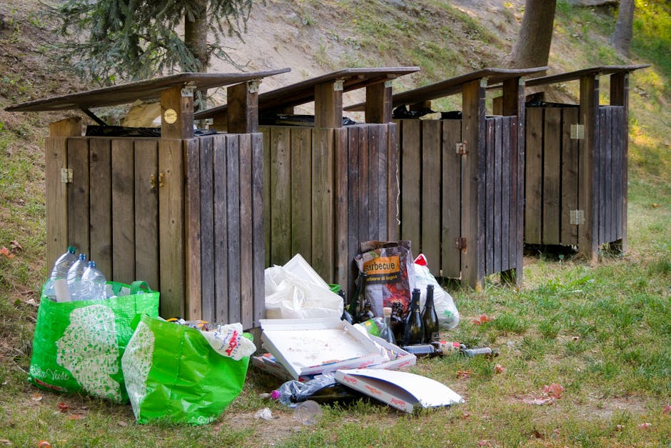 A vertical view of the exterior side of a green wooden building with weathered, textured cladding. A set of wooden stairs with a brown finish leads to a small white entrance door, positioned just above a concrete sidewalk. Several black garbage bags, filled and tied, are piled against the building near the base of the stairs, occupying part of the pavement. To the left, a partially locked metal door with graffiti and peeling paint is visible, along with a small white utility box attached to the wall. In the background, an urban street scene includes parked cars, a large brick building, and power lines overhead under a cloudy sky. The scene reflects a typical environment where independent rubbish collection and waste management services, like those offered by Rubbish Removal Kensington, might be used to handle overflowing refuse or bulk waste outside residential buildings.