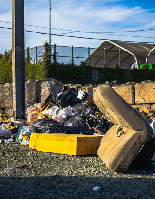 A large collection of overflowing rubbish bins and scattered waste, including black, red, and gray bins, with some lids open to reveal mixed paper, cardboard, plastic bags, and various general household waste. The waste is piled on the pavement and partially pushed out of the bins, with some items spilling onto the ground. Behind the bins, there is a metal railing and a parked silver car with a license plate reading 'H1 OBE,' positioned on a cobbled street. In the background, a two-story building features blue scaffolding and protective netting on the upper level, with visible signage indicating commercial spaces such as a fish bar. The scene appears to be an area where skip or bin collections have been delayed or missed, illustrating the need for professional rubbish removal services like those offered by Rubbish Removal Kensington, especially for private waste collection or on-site clearance in urban settings.