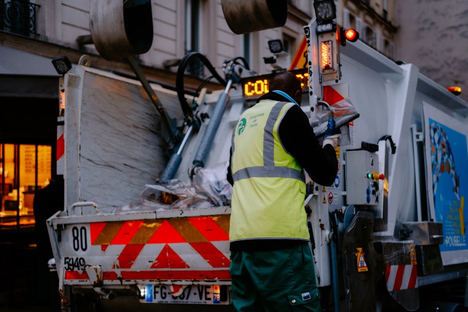 A worker wearing a high-visibility yellow vest and dark clothing is operating a white waste collection truck in an urban street setting during dusk or early evening. The worker, seen from the back, is adjusting controls on the side of the truck, which features reflective orange and red safety markings. The truck is loaded with accumulated rubbish, including large, irregularly shaped piles of mixed waste covered with white and gray materials, possibly plastic or paper. The environment shows a building with illuminated windows in the background, adding a warm glow to the scene. The lighting suggests natural twilight combined with street or building lights, emphasizing the operational context of independent waste collection and rubbish removal services conducted by Rubbish Removal Kensington. The overall scene underscores the practical process of on-site waste handling in an urban commercial or residential area, with the worker actively engaged in maintaining efficient rubbish collection, supporting alternative disposal methods outside traditional local authority services.