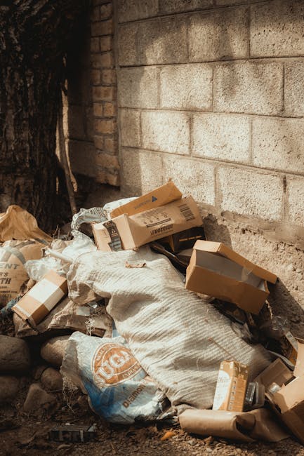 A collection of various discarded cardboard boxes, some flattened and others partially intact, piled against a rough-textured brick wall in an outdoor setting. The boxes are of different sizes, with brown and white surfaces, some featuring printed labels or barcodes. Among the boxes, there are also plastic bags filled with rubbish, including a large woven sack and smaller not clearly identifiable bags. The ground beneath the rubbish appears to be dirt and small rocks, and a section of a tree trunk is visible to the left side of the image. The scene suggests an area of private waste collection or an on-site clearance, provided by a waste removal service such as Rubbish Removal Kensington. The lighting is natural, with shadows cast by nearby objects, emphasizing the cluttered nature of this waste disposal scene against the backdrop of a brick wall, indicative of a rear garden or alleyway involved in domestic waste management activities.