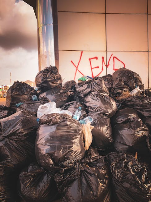 A worker wearing a high-visibility yellow vest and dark clothing is operating a white waste collection truck in an urban street setting during dusk or early evening. The worker, seen from the back, is adjusting controls on the side of the truck, which features reflective orange and red safety markings. The truck is loaded with accumulated rubbish, including large, irregularly shaped piles of mixed waste covered with white and gray materials, possibly plastic or paper. The environment shows a building with illuminated windows in the background, adding a warm glow to the scene. The lighting suggests natural twilight combined with street or building lights, emphasizing the operational context of independent waste collection and rubbish removal services conducted by Rubbish Removal Kensington. The overall scene underscores the practical process of on-site waste handling in an urban commercial or residential area, with the worker actively engaged in maintaining efficient rubbish collection, supporting alternative disposal methods outside traditional local authority services.