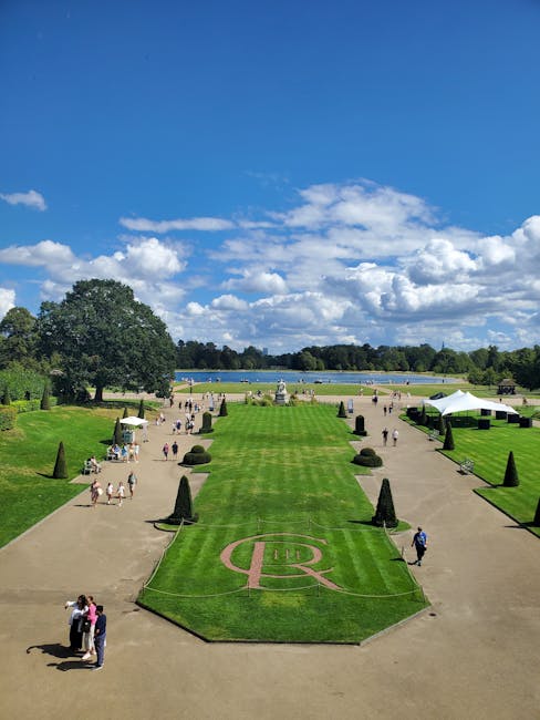 A wide view of a formal park landscape featuring neatly maintained green lawns and pathways, with several groups of people walking and gathering. In the foreground, a grass area displays a large letter 'R' within a circle, bordered by shaped shrubs and conifer trees aligned along the paths. To the right, a white tent and some outdoor furniture are set up near the walking area. In the background, tall trees and hedges frame a large lake or reservoir, with a clear blue sky overhead dotted with fluffy white clouds. The scene suggests a well-organized outdoor space that may be used for public events or private gatherings, with no visible rubbish or debris, but the description aligns with contexts where independent waste clearance services like Rubbish Removal Kensington could be involved in maintaining public or private landscaped spaces.