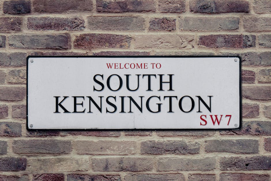 A rectangular metal street sign mounted on a light brown brick wall, displaying the text 'Welcome to South Kensington SW7' in bold black letters, with 'Welcome to' and 'SW7' in smaller red font. The sign features a smooth, white background and slightly rounded corners, with visible screws at each corner securing it to the wall. The brick wall behind the sign consists of standard-sized bricks with a mottled texture, warm tones of beige and reddish-brown, and visible mortar lines. The lighting appears natural and evenly distributed, illuminating the sign clearly and casting minimal shadows. This scene suggests an urban or residential area in South Kensington, typical of private or public spaces where such signage indicates a notable district. The clean and orderly appearance of the sign and wall contribute to an overall neutral and professional visual impression, aligning with the context of property or district identification relevant to waste management and local infrastructure services.