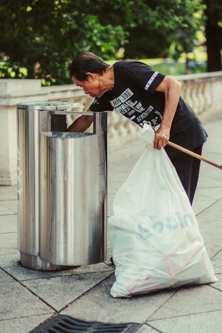 A man with short dark hair, wearing a black T-shirt with white and blue text, is seen disposing of waste into a stainless steel litter bin on a sidewalk. He is holding a large white trash bag in his left hand and using his right hand to reach into the open-top bin. The bin is positioned against a low stone wall, with a background of lush green trees and foliage, indicating an outdoor urban environment. The pavement is paved with large square tiles and a drain is visible in the foreground. The scene depicts an urban setting where individual waste disposal is occurring, aligning with private rubbish removal or alternative waste handling services such as those offered by Rubbish Removal Kensington in South Kensington, SW7. The lighting appears natural, suggesting daytime, with soft shadows cast by the trees and the man’s figure.