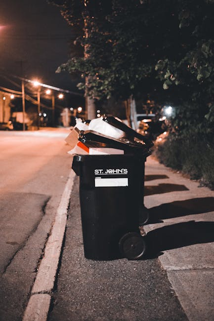 A black wheeled rubbish bin positioned on a pavement beside a narrow street during nighttime, with a white label on the front indicating it belongs to St. John's. The bin's open lid reveals mixed waste, including paper and cardboard. The street is lined with trees and bushes, with streetlights casting warm, orange glows onto the road and sidewalk. In the background, faint outlines of parked cars and illuminated buildings suggest an urban residential area. The scene highlights an example of private waste disposal, possibly provided by a local rubbish removal service like Rubbish Removal Kensington, for collecting household waste and bulky items not suitable for standard rubbish collection. The lighting and environment suggest a typical city street with limited space, illustrating the need for individual rubbish management solutions for residents in narrow Kensington streets, especially for bulky waste that requires special access or separate collection arrangements.