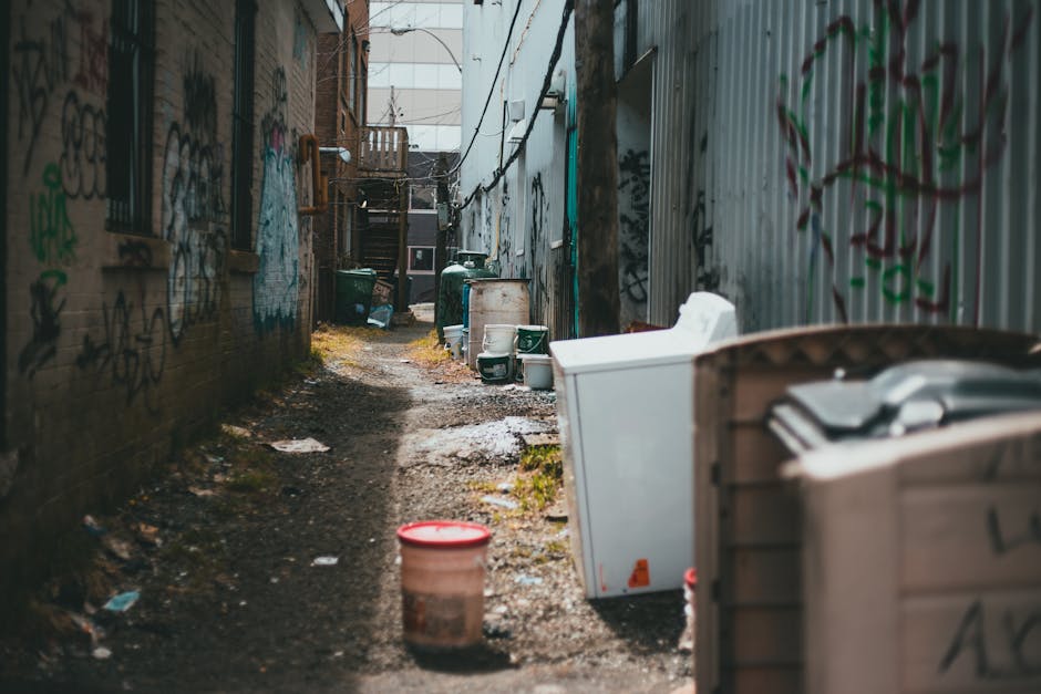 A narrow outdoor alleyway during daytime, with a dirt and gravel surface showing patches of moss and small puddles. On the left side, a brick wall covered in multicoloured graffiti extends into the distance, with a small window secured by metal bars visible partway along. On the right side, a corrugated metal fence, also marked with graffiti, runs parallel to the brick wall, creating a confined space. Various discarded objects are placed along both sides of the alley, including several large white appliances like washing machines and refrigerators leaning against the fence, some partially obscured and appearing weathered. In the foreground, a plastic bucket with a red lid sits on the ground, slightly muddy and dirty. Further back, green waste bins are lined up near the end of the alley, with a few smaller items and pieces of debris scattered around. The scene appears to depict an area used for private waste disposal or irregular rubbish collection, with the environment showing signs of neglect typical in locations managed by independent rubbish removal services like Rubbish Removal Kensington, serving narrow urban streets.
