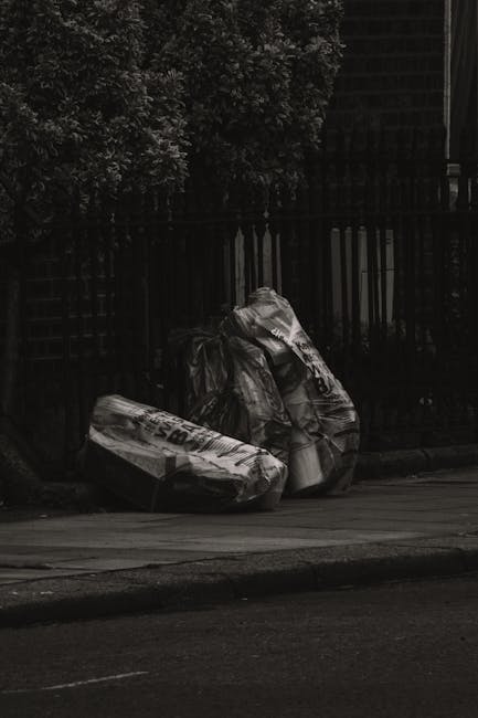 Two large black plastic rubbish bags, appearing crumpled and somewhat weathered, are placed on the sidewalk next to a low curb, with a few smaller bags visible nearby. The bags are filled with waste and are leaning slightly against a black metal fence, which features vertical bars with decorative top elements. Behind the fence, dense foliage from a large bush or small tree can be seen, with dark, leafy branches contrasting against the darker background. The surrounding environment suggests an urban residential area, with a paved pavement and a road surface in the foreground. The scene is outdoor and appears to be during the daytime, under natural lighting, with no visible activity or individuals present. The setting emphasizes the need for effective rubbish disposal or alternative waste handling, such as private rubbish collection by Rubbish Removal Kensington, as evidenced by the presence of waste accumulated outside standard collection times.