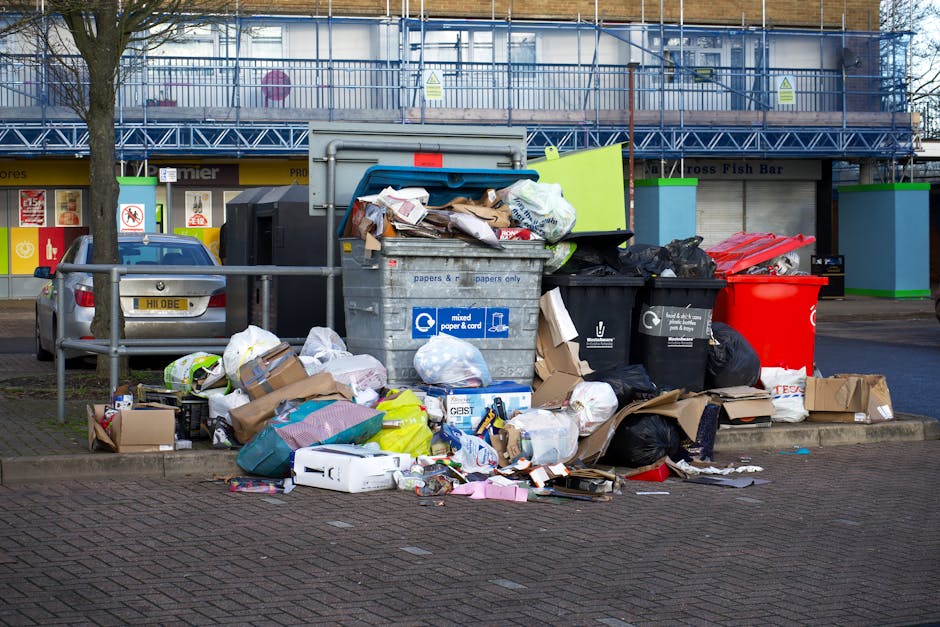 A pile of mixed refuse consisting of flattened cardboard boxes, paper bags, and crumpled packaging material, piled against a rough, uneven brick and concrete wall in an outdoor setting. The cardboard boxes vary in size and color, with some opened or partially collapsed. A large white sack made of woven material is partially visible, containing or supporting some of the debris, and appears weathered with dirt and slight staining. The surrounding ground is composed of small rocks and dirt, with a tree trunk visible on the left side of the image. The lighting suggests natural daylight, creating subtle shadows across the scene. This scene illustrates a common example of waste accumulation typical of non-regular rubbish collection, highlighting the types of materials that might be handled during private waste removal services, such as those provided by Rubbish Removal Kensington, for on-site clearance or alternative waste handling outside of municipal rubbish collection schedules.